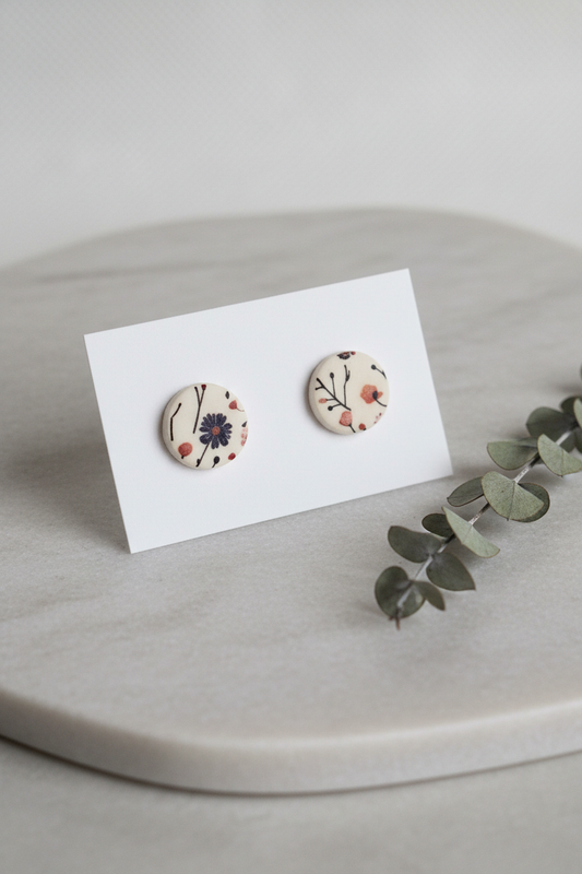 Handmade polymer clay Wild Stem circle stud earrings — sparse botanical illustration print with wispy stems and seed heads on cream, stainless steel posts, on white jewellery card on stone surface with dried seed head