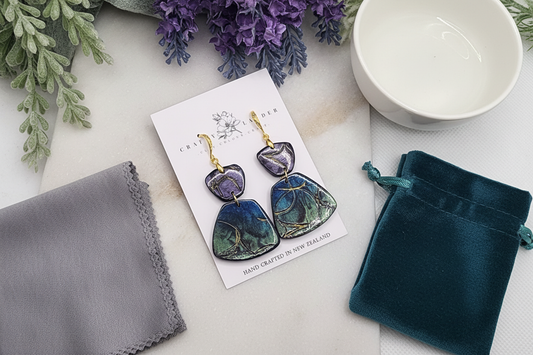 "Jewelry care flat-lay showing Crafty Larder handcrafted statement earrings with iridescent marbled pendants, microfiber cleaning cloth, water bowl, and storage pouch"