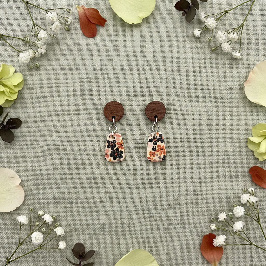 Overhead flat lay of Crafty Larder Autumn Meadow earrings on sage green linen with scattered petals — round dark wood stud with small floral trapezoid clay drop in warm rust and navy Liberty print on cream