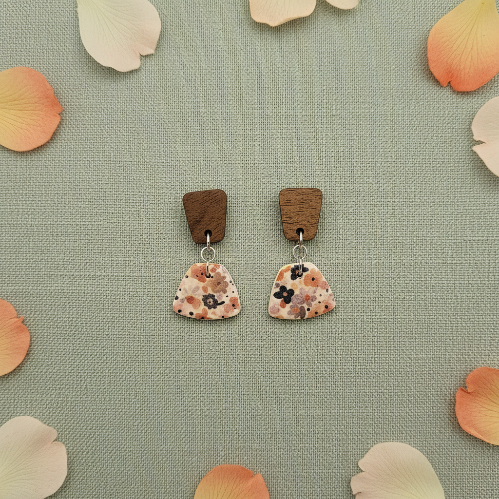 Overhead flat lay of Crafty Larder Autumn Meadow earrings on sage green linen with scattered petals — dark wood square stud with small floral fan clay drop in warm rust, peach and navy Liberty print on cream