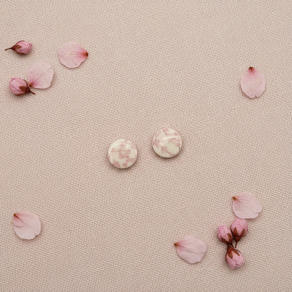 Overhead flat lay of Crafty Larder Blossom Branch earrings on blush pink linen with dried cherry blossom petals — small round polymer clay circle studs with delicate soft pink cherry blossom print on cream