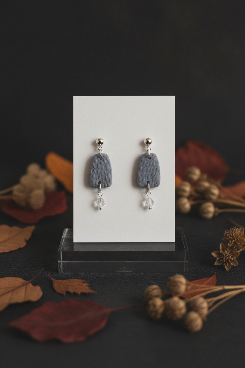 Handmade polymer clay Botanical Crystal Earrings in Vapor Blue — soft grey-blue rounded trapezoid textured drop with silver ball stud top, silver hoop connector and delicate crystal bead dangle, displayed on a white jewellery card with moody autumn botanical props, handcrafted in New Zealand by Crafty Larder