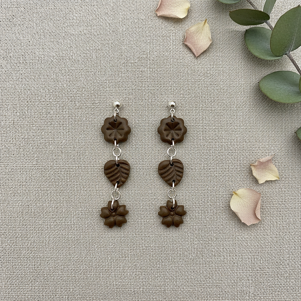 Flat lay of Crafty Larder Botanical Trio Earrings in Bronze Brown on warm grey linen with eucalyptus — handmade polymer clay three-tier flower, leaf & flower drop earrings in deep olive-brown