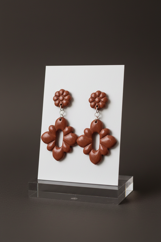 Handmade polymer clay Gerbera Drop Earrings in Chili Oil — warm terracotta brown gerbera flower stud top with quatrefoil drop, displayed on a plain white jewellery card on a clear acrylic stand