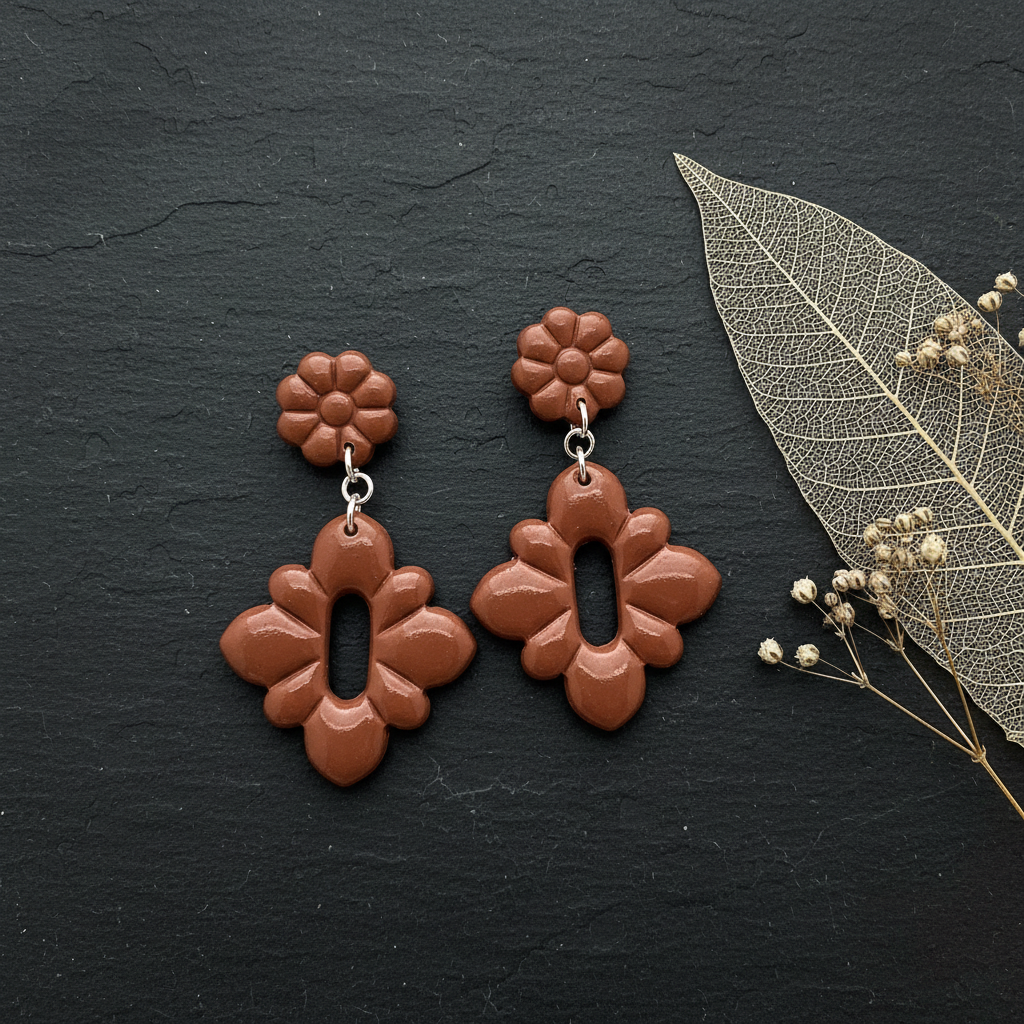 Flat lay of Crafty Larder Gerbera Drop Earrings in Chili Oil on a dark moody surface with dried botanicals — handmade polymer clay dangle earrings in warm terracotta brown