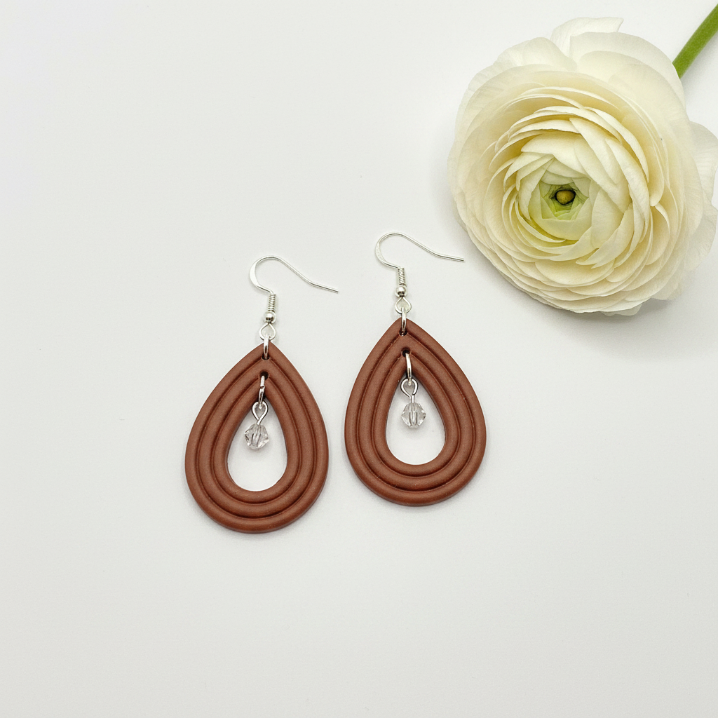 Overhead flat lay of Crafty Larder Raindrop Crystal Earrings in Chili Oil on white surface with fresh flower — handmade polymer clay open teardrop hoop earrings with crystal bead drop in warm medium brown