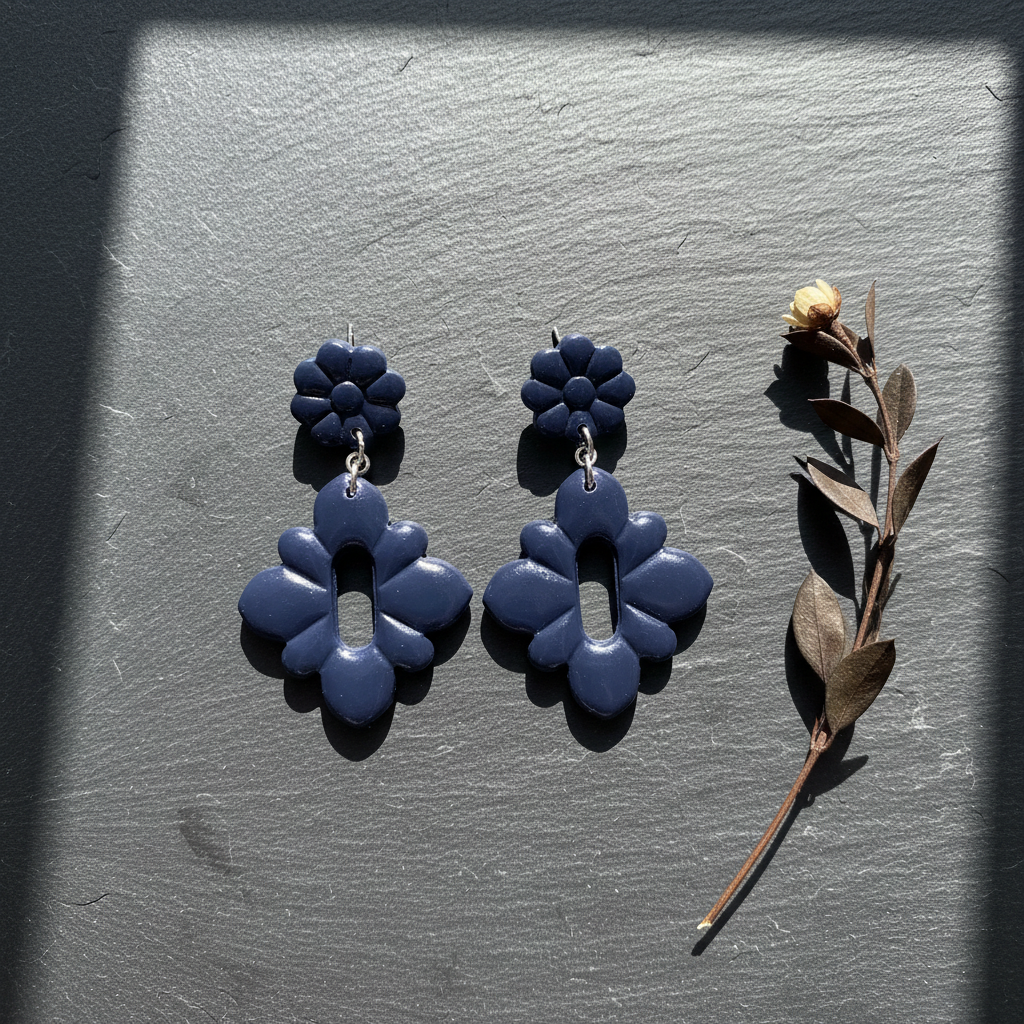Flat lay of Crafty Larder Gerbera Drop Earrings in Crown Blue on a dark moody surface with dried botanicals — handmade polymer clay dangle earrings in deep midnight navy