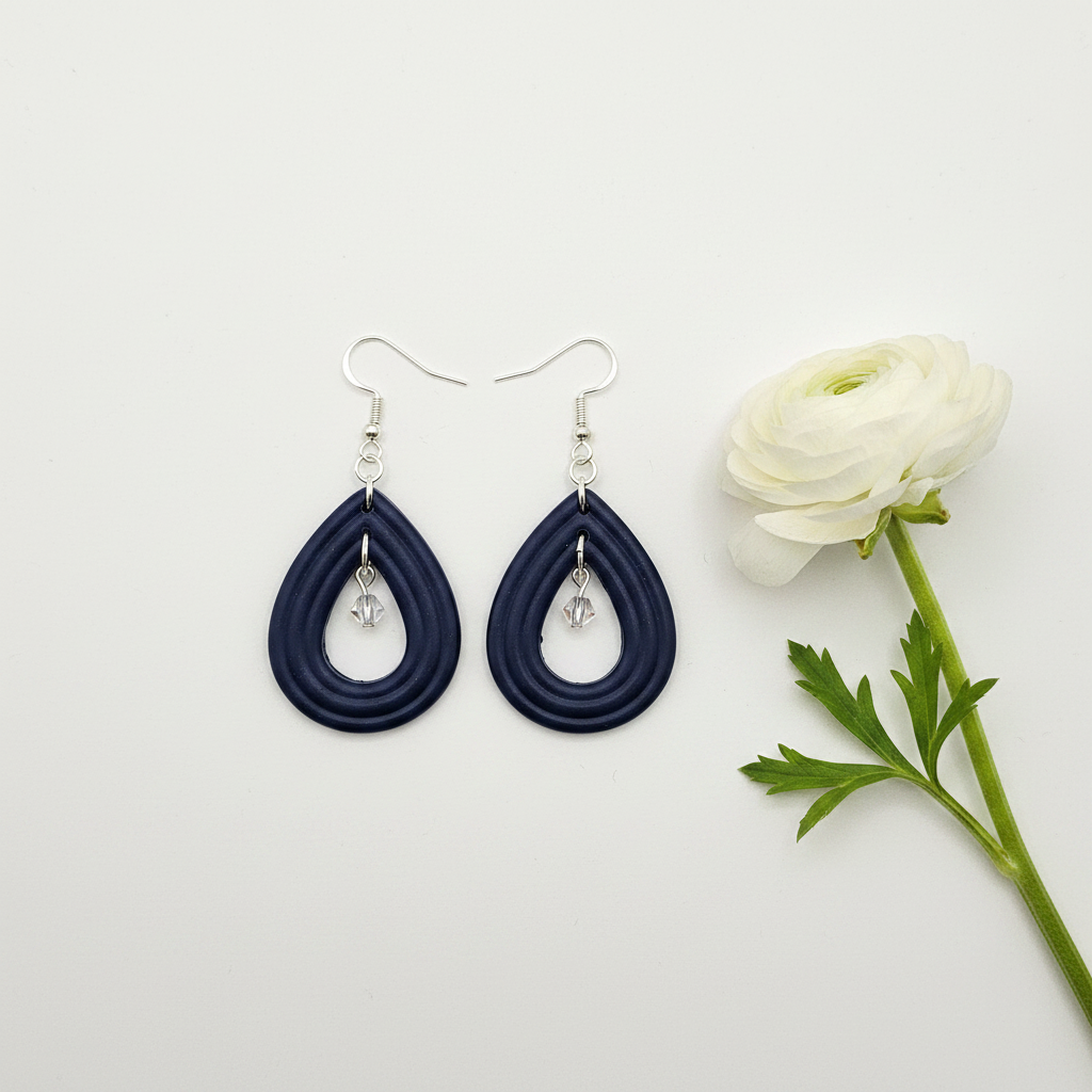 Overhead flat lay of Crafty Larder Raindrop Crystal Earrings in Crown Blue on white surface with fresh flower — handmade polymer clay open teardrop hoop earrings with crystal bead drop in deep midnight navy