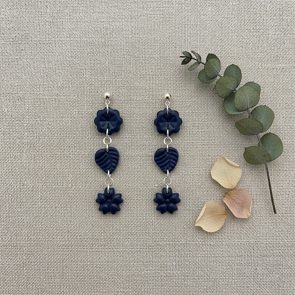 Flat lay of Crafty Larder Botanical Trio Earrings in Crown Blue on warm grey linen with eucalyptus — handmade polymer clay three-tier flower, leaf & flower drop earrings in deep midnight navy