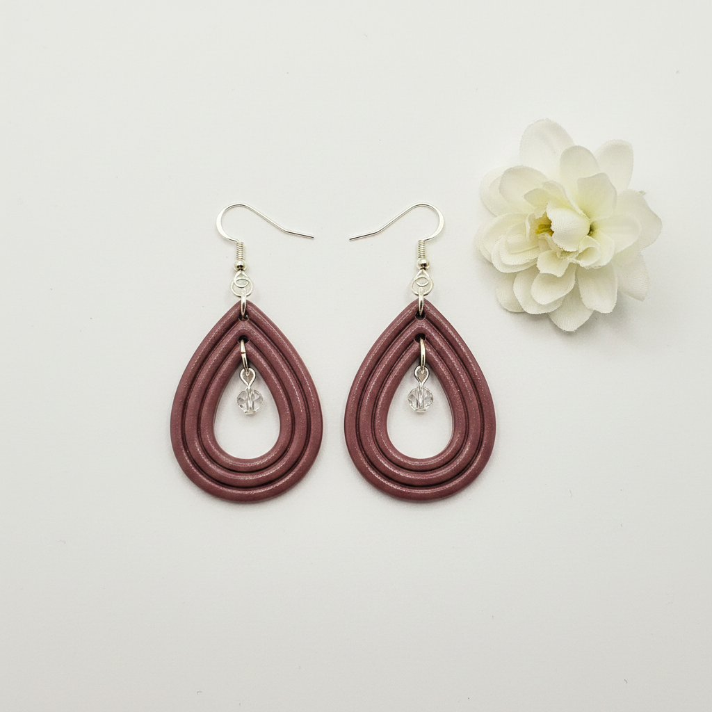 Overhead flat lay of Crafty Larder Raindrop Crystal Earrings in Damson on white surface with fresh flower — handmade polymer clay open teardrop hoop earrings with crystal bead drop in muted dusty plum-mauve