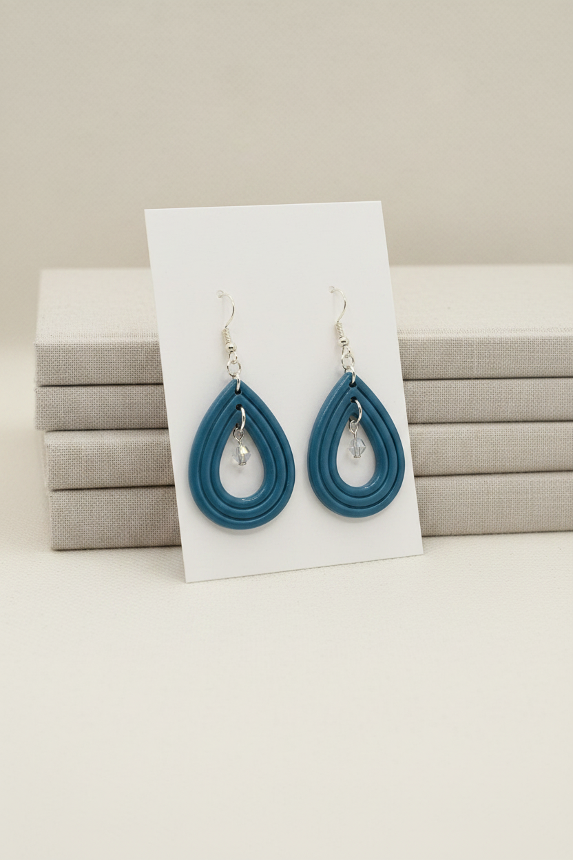 andmade polymer clay Raindrop Crystal Earrings in Lyons Blue — muted teal-blue open teardrop hoop earrings with crystal bead drop, displayed on a plain white jewellery card propped against linen