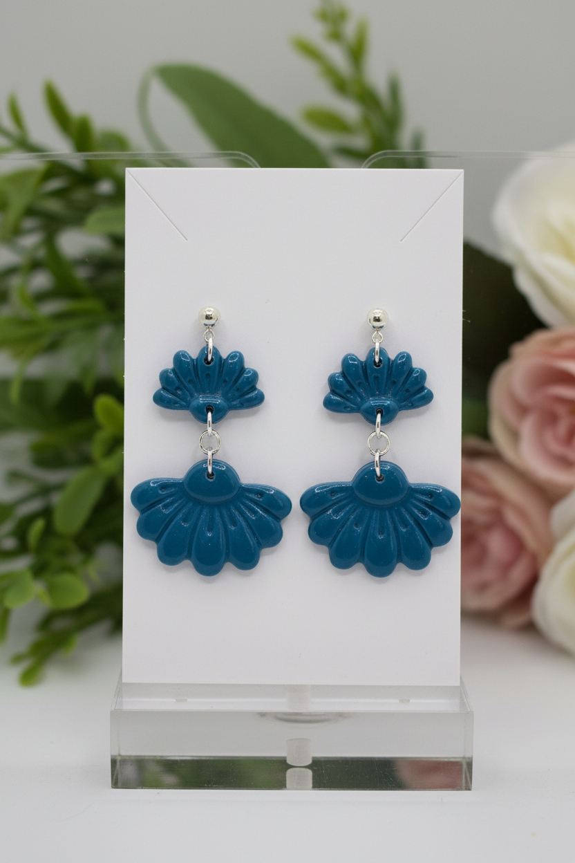 Handmade polymer clay Petal Tier Earrings in Lyons Blue — teal-blue two-tiered fan-shaped ruffled drops with silver ball stud, displayed on a plain white jewellery card with soft botanical background
