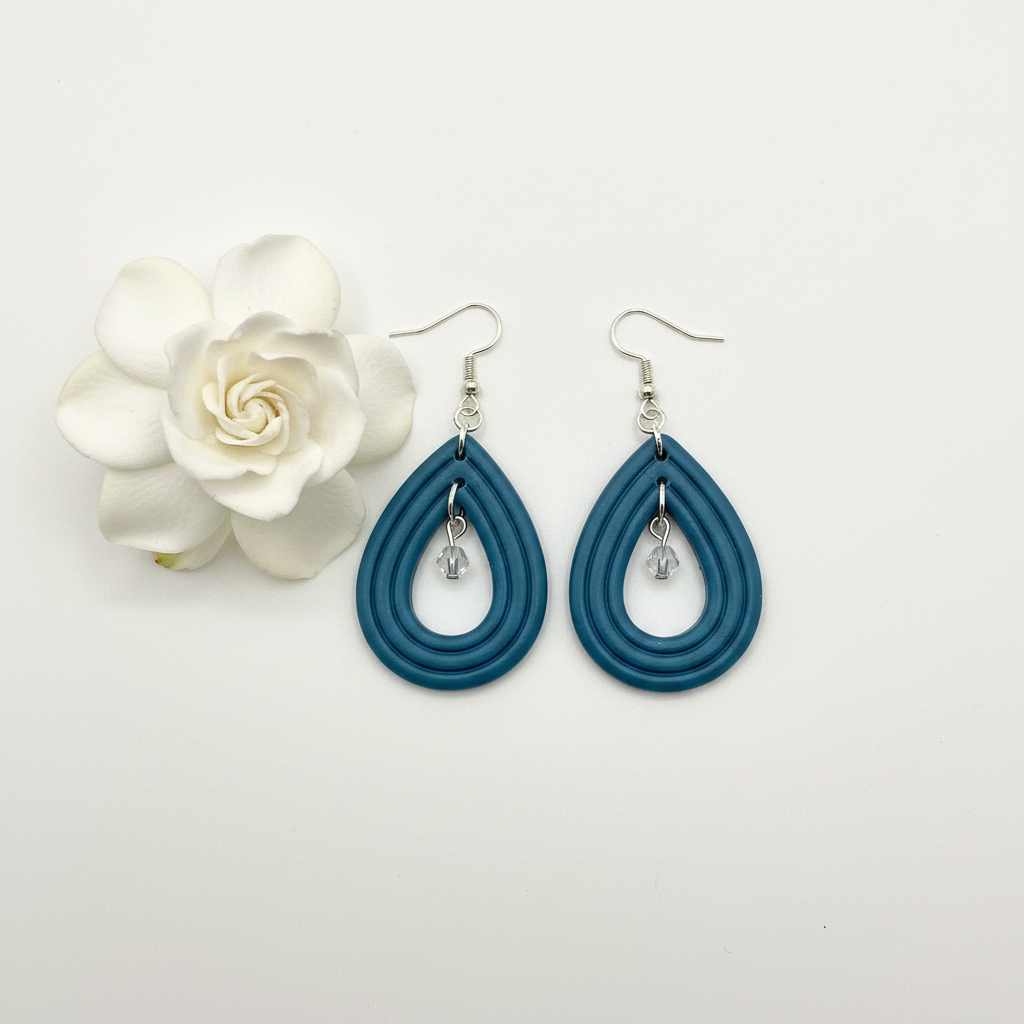 Overhead flat lay of Crafty Larder Raindrop Crystal Earrings in Lyons Blue on white surface with fresh flower — handmade polymer clay open teardrop hoop earrings with crystal bead drop in muted teal-blue