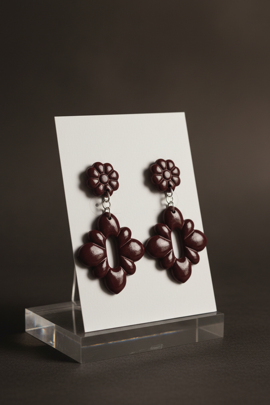 Handmade polymer clay Gerbera Drop Earrings in Mauve Wine — deep burgundy gerbera flower stud top with quatrefoil drop, displayed on a plain white jewellery card on a clear acrylic stand