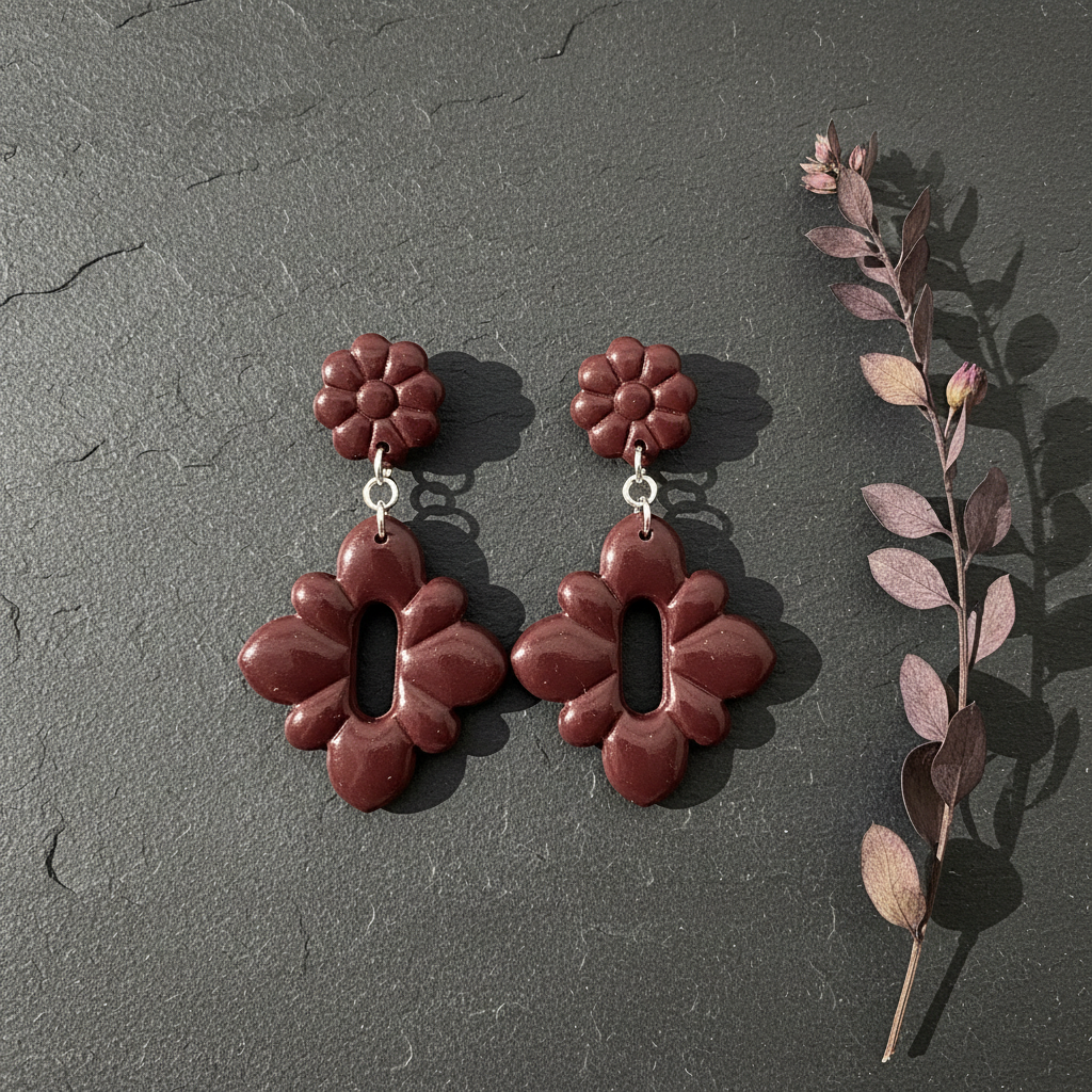 Flat lay of Crafty Larder Gerbera Drop Earrings in Mauve Wine on a dark moody surface with dried botanicals — handmade polymer clay dangle earrings in deep burgundy
