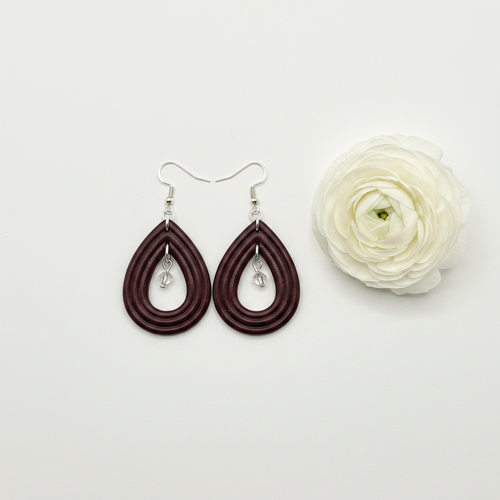 Overhead flat lay of Crafty Larder Raindrop Crystal Earrings in Mauve Wine on white surface with fresh flower — handmade polymer clay open teardrop hoop earrings with crystal bead drop in deep dark burgundy