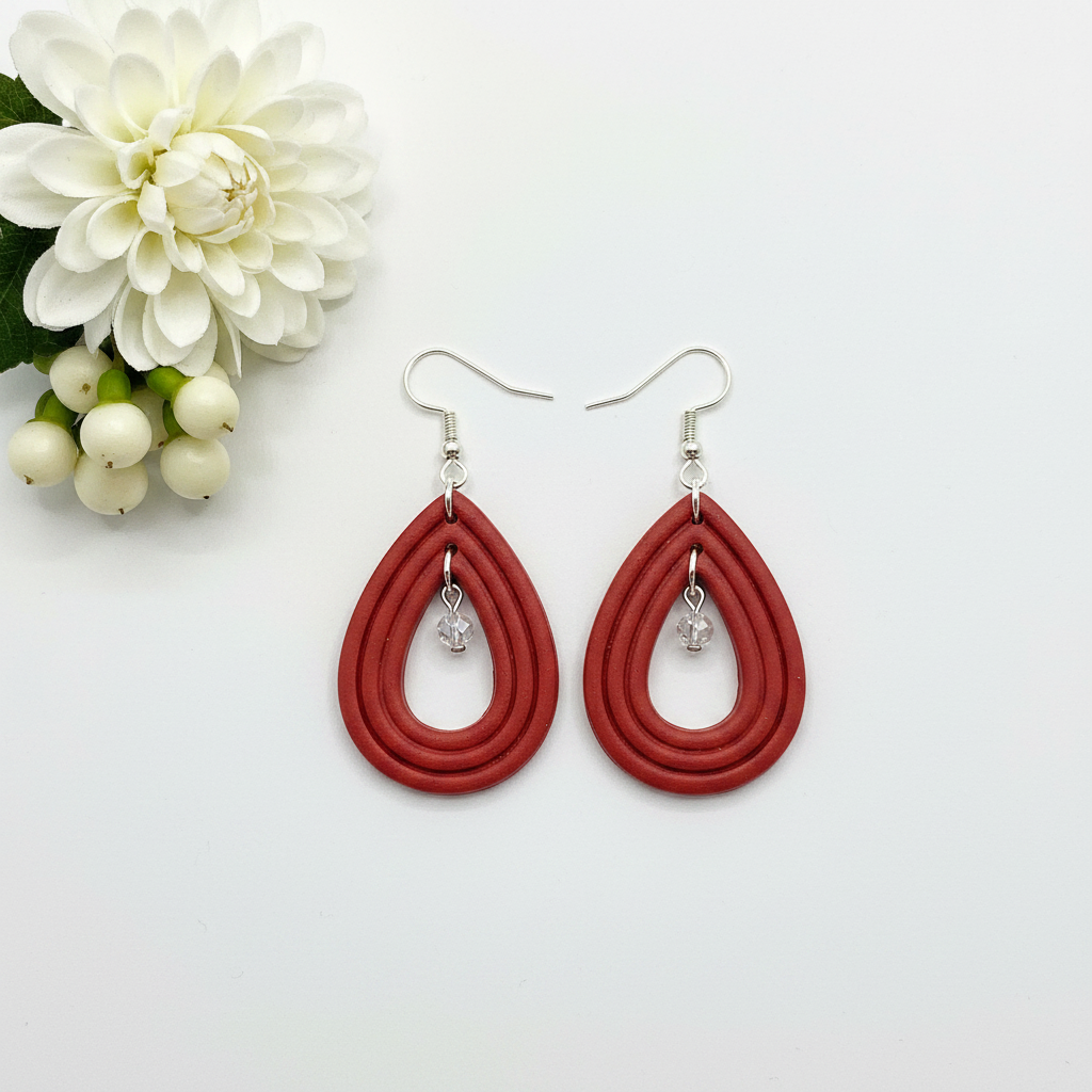Overhead flat lay of Crafty Larder Raindrop Crystal Earrings in Poppy Red on white surface with fresh flower — handmade polymer clay open teardrop hoop earrings with crystal bead drop in warm brick red
