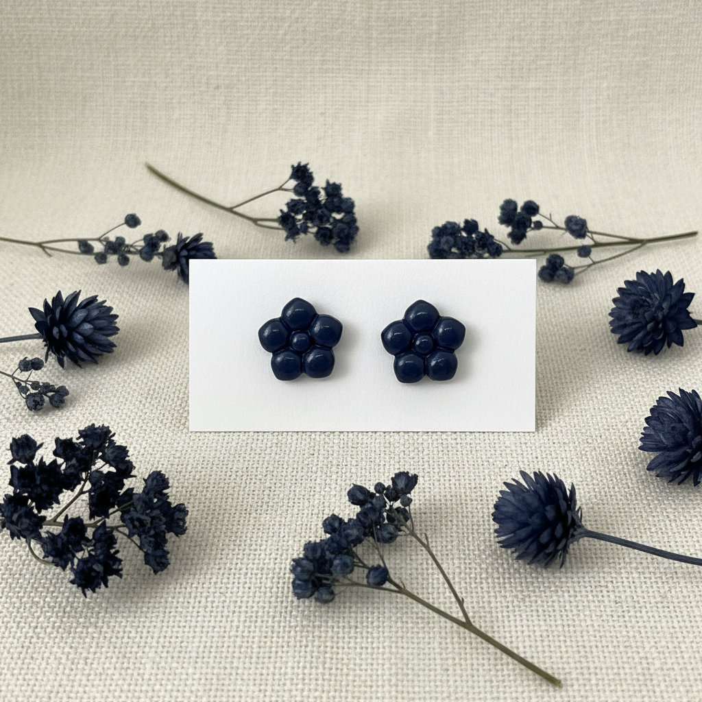 "Primrose Studs in Crown Blue — handmade polymer clay flower stud earrings with smooth glossy bubble petal finish, displayed on white card with deep navy dried botanicals and dark indigo botanical styling, handcrafted in New Zealand by Crafty Larder