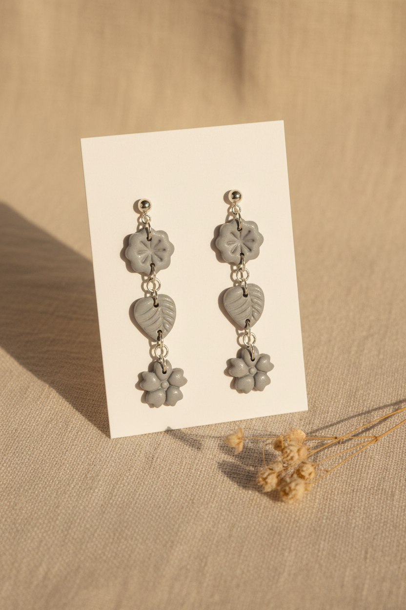 Handmade polymer clay Botanical Trio Earrings in Vapor Blue — soft cool grey-blue three-tier flower, leaf & flower drop earrings on a plain white jewellery card with dried botanical stem