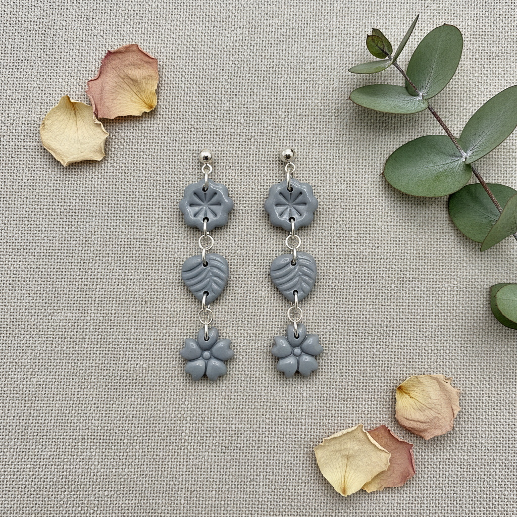 Flat lay of Crafty Larder Botanical Trio Earrings in Vapor Blue on warm grey linen with eucalyptus — handmade polymer clay three-tier flower, leaf & flower drop earrings