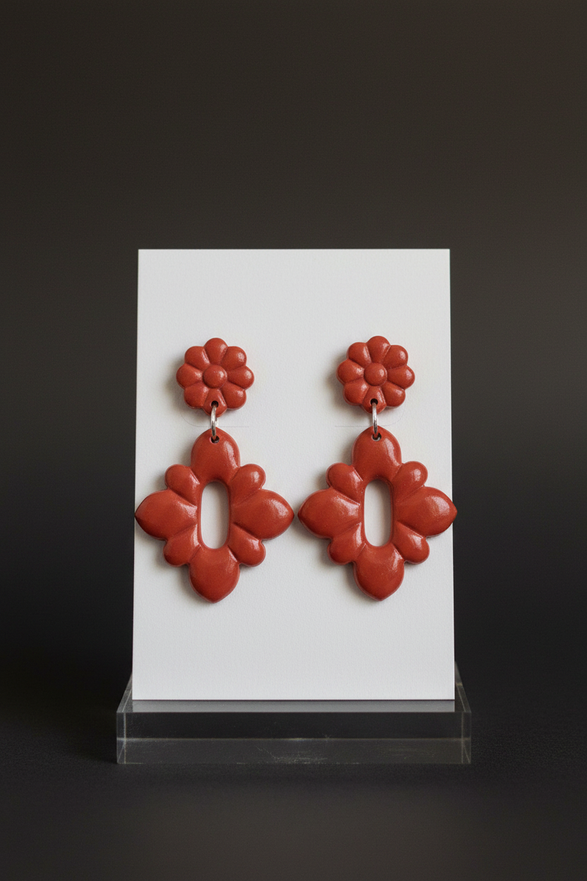 Handmade polymer clay Gerbera Drop Earrings in Winterberry — warm terracotta red gerbera flower stud top with quatrefoil drop, displayed on a plain white jewellery card on a clear acrylic stand