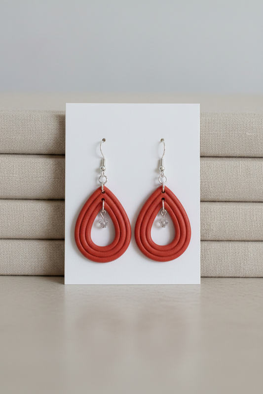 Handmade polymer clay Raindrop Crystal Earrings in Winterberry — warm terracotta red open teardrop hoop earrings with crystal bead drop, displayed on a plain white jewellery card propped against linen