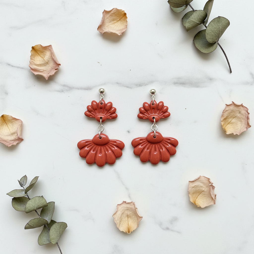 Flat lay of Crafty Larder Petal Tier Earrings in Winterberry on white marble with scattered petals — handmade polymer clay two-tiered ruffled drop earrings in warm terracotta red