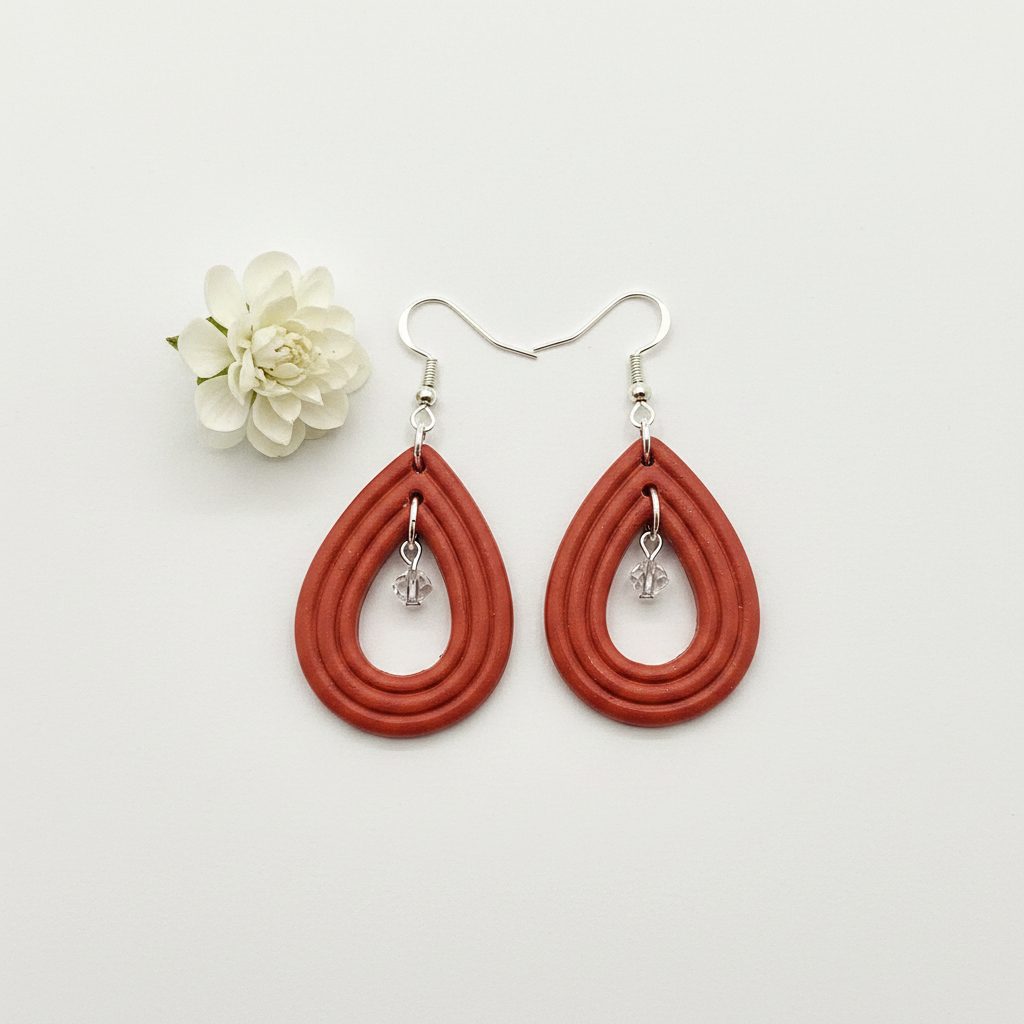 Overhead flat lay of Crafty Larder Raindrop Crystal Earrings in Winterberry on white surface with fresh flower — handmade polymer clay open teardrop hoop earrings with crystal bead drop in warm terracotta red